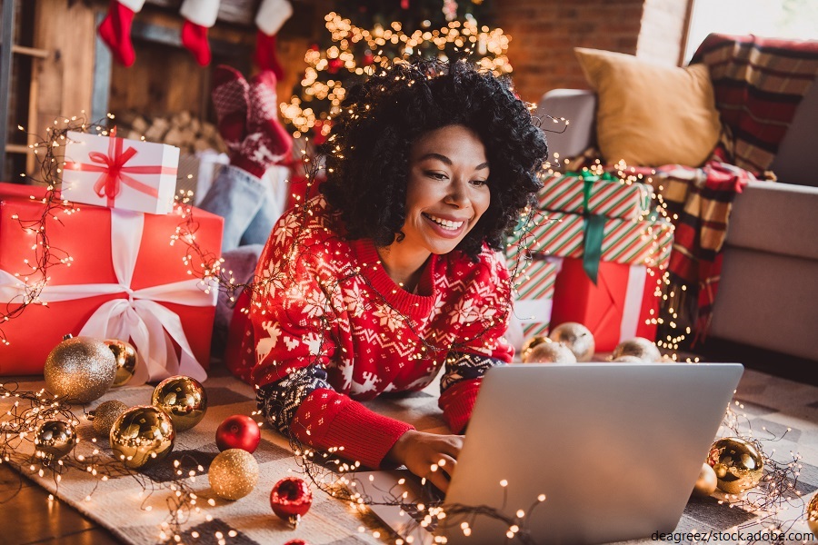 Frau liegt auf dem Boden vor ihrem Laptop umgeben von weihnachtlichen Accessoires und Geschenken